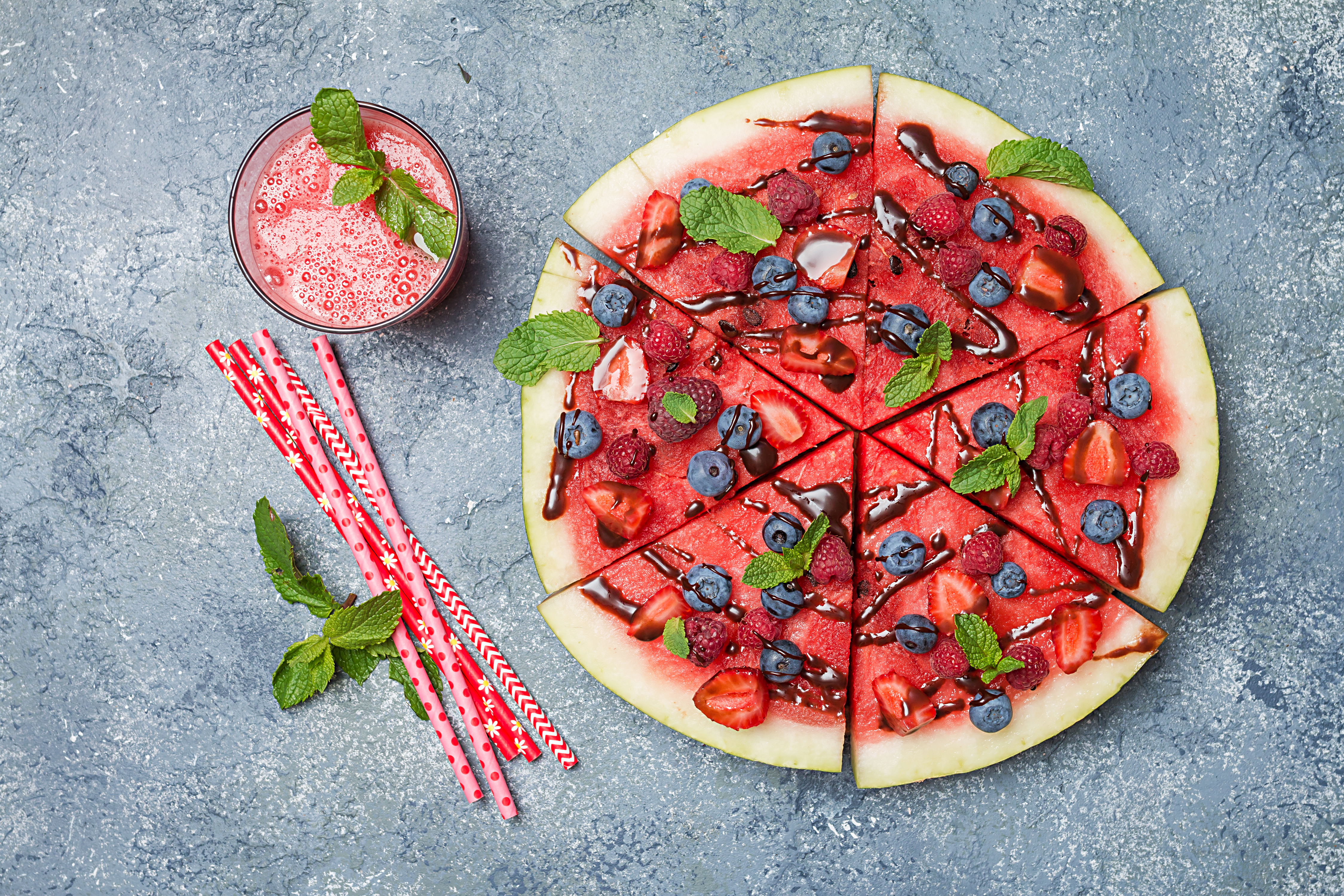 colorful tropical fruit watermelon pizza topped with berries and chocolate sauce with watermelon juice over gray stone background, top view