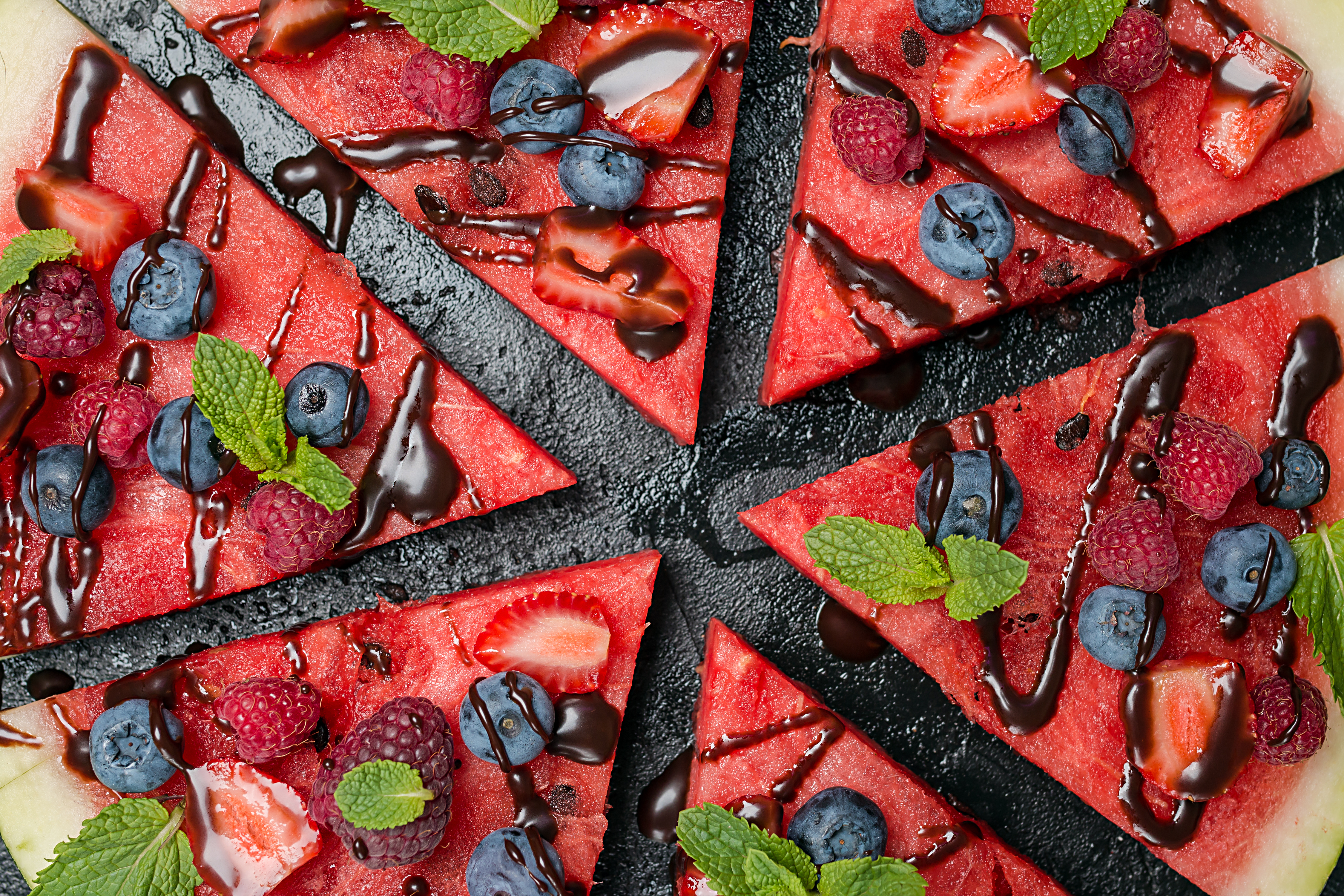 colorful tropical fruit watermelon pizza topped with berries and chocolate sauce cut into segments over black background, top view